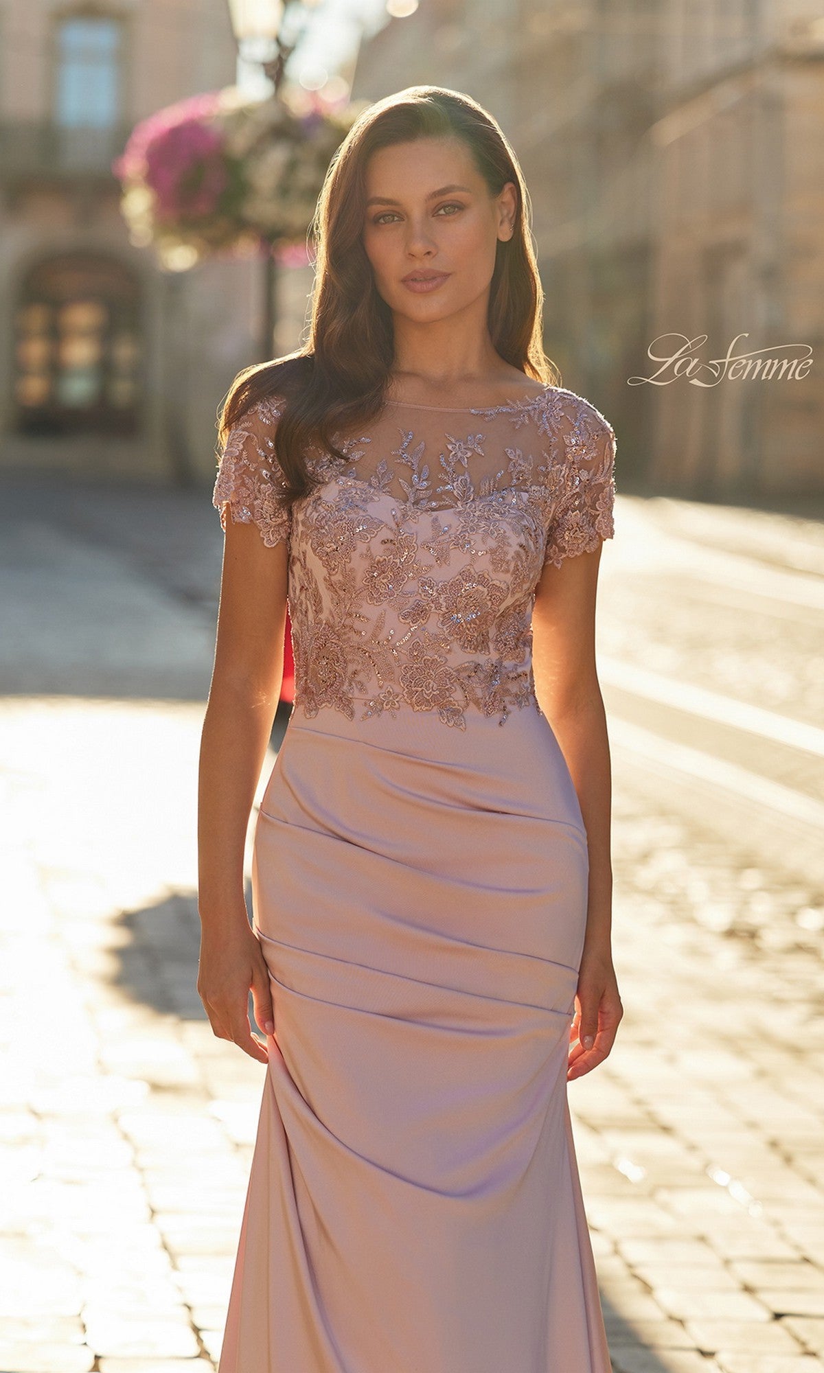 A woman stands outdoors in soft sunlight, wearing the La Femme 32737 long formal dress—light pink with an illusion neckline, lace embroidered bodice, short sleeves, and a flowing skirt.